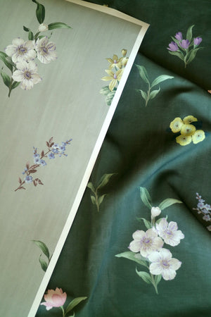 A close-up of floral wallpaper or fabric with a dark green background, featuring white cherry blossoms, yellow flowers, and purple sprigs. A rectangular sample card showing the pattern is placed diagonally in the foreground.