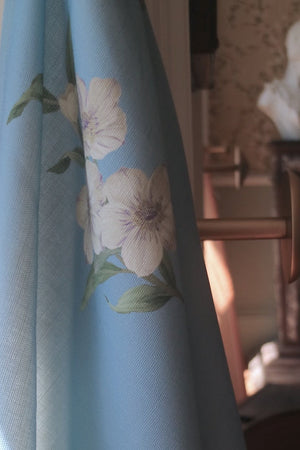 A light blue curtain with a delicate floral print hanging in a sunlit room next to a dark wicker basket and a classical bust statue in the background.